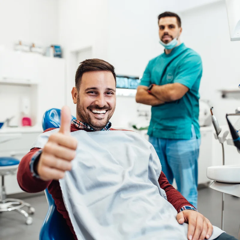 Male patient giving thumbs up in dental chair after successful treatment with dentist in background