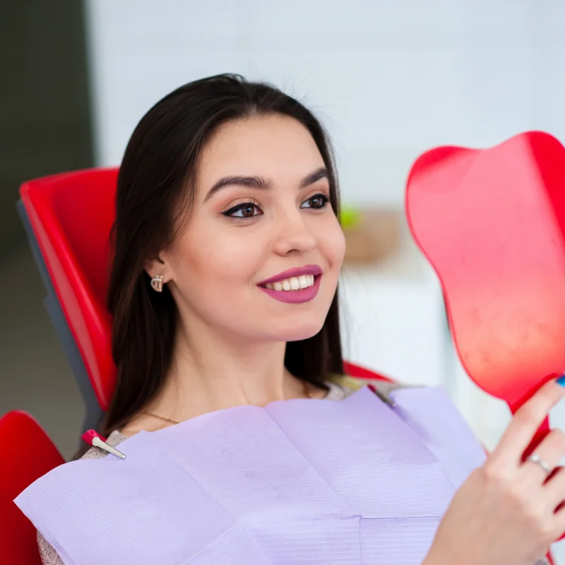 Female patient in dental chair checking smile in mirror after cosmetic dental treatment