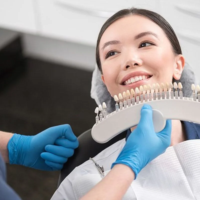 Dentist holding veneer shade guide next to female patient's teeth during veneer consultation