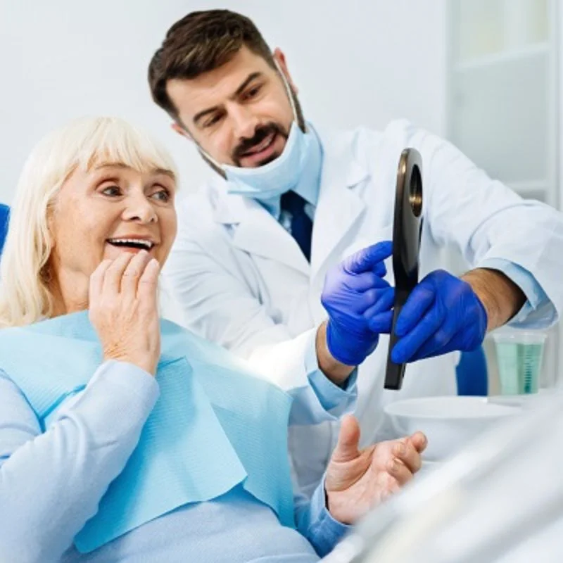 Elderly female patient smiling with surprise as dentist shows her new dentures in a mirror
